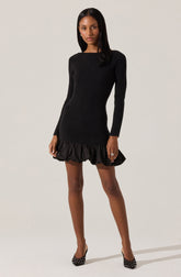 A woman is wearing a fitted, long-sleeved black ribbed knit dress with a ruffled hemline. She is also wearing black high heels with white polka dots.
